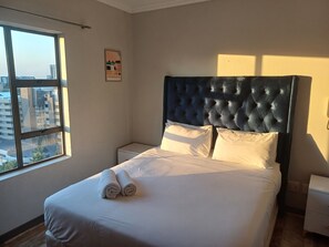 Ein ordentlich bezogenes Bett mit weißen Leinen und aufgerollten Handtüchern, ein dunkel gestuftes Kopfteil, ein kleiner Nachttisch und ein Fenster mit Blick auf Gebäude.