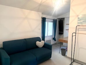 Ein Zimmer mit einem dunkelgrünen Sofa, einem herzförmigen Kissen, einem Fenster mit blauen Vorhängen, einem Holztisch und einem schwarzen Metallregal.