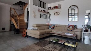 A living room with a sofa, coffee table, and a guitar. There is a staircase and a window with a view. - A l'ombre des 2 tilleuls (Sainte-Pazanne)