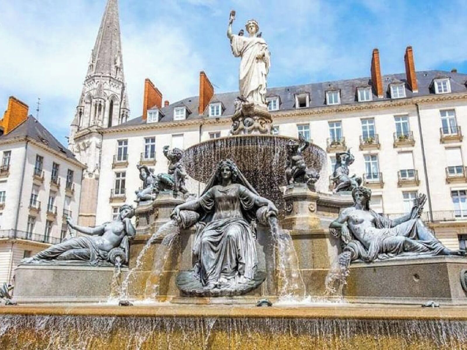 Une fontaine ornée de statues, un bâtiment avec de nombreuses fenêtres et une tour en arrière-plan.