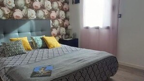 A bedroom with a bed, patterned bedding, a headboard, a nightstand, and a wall with a floral wallpaper. - A l'ombre des 2 tilleuls (Sainte-Pazanne)