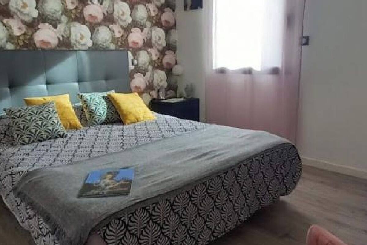 A bedroom with a bed, patterned bedding, a headboard, a nightstand, and a wall with a floral wallpaper.