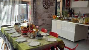 A dining area with a long table set for six, a kitchen in the background, and a brick wall. - A l'ombre des 2 tilleuls (Sainte-Pazanne)