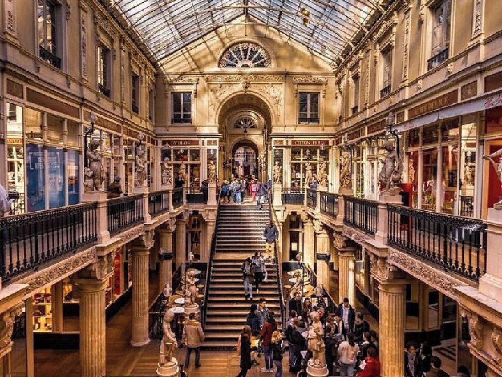 Galería comercial de varios niveles con arquitectura ornamentada, que incluye columnas y estatuas, y una escalera central.