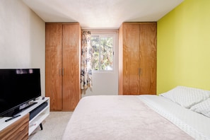 Ein Schlafzimmer mit einem Bett, einem Fernseher auf einem Schrank, Holzschränken und einem Fenster mit Vorhängen.