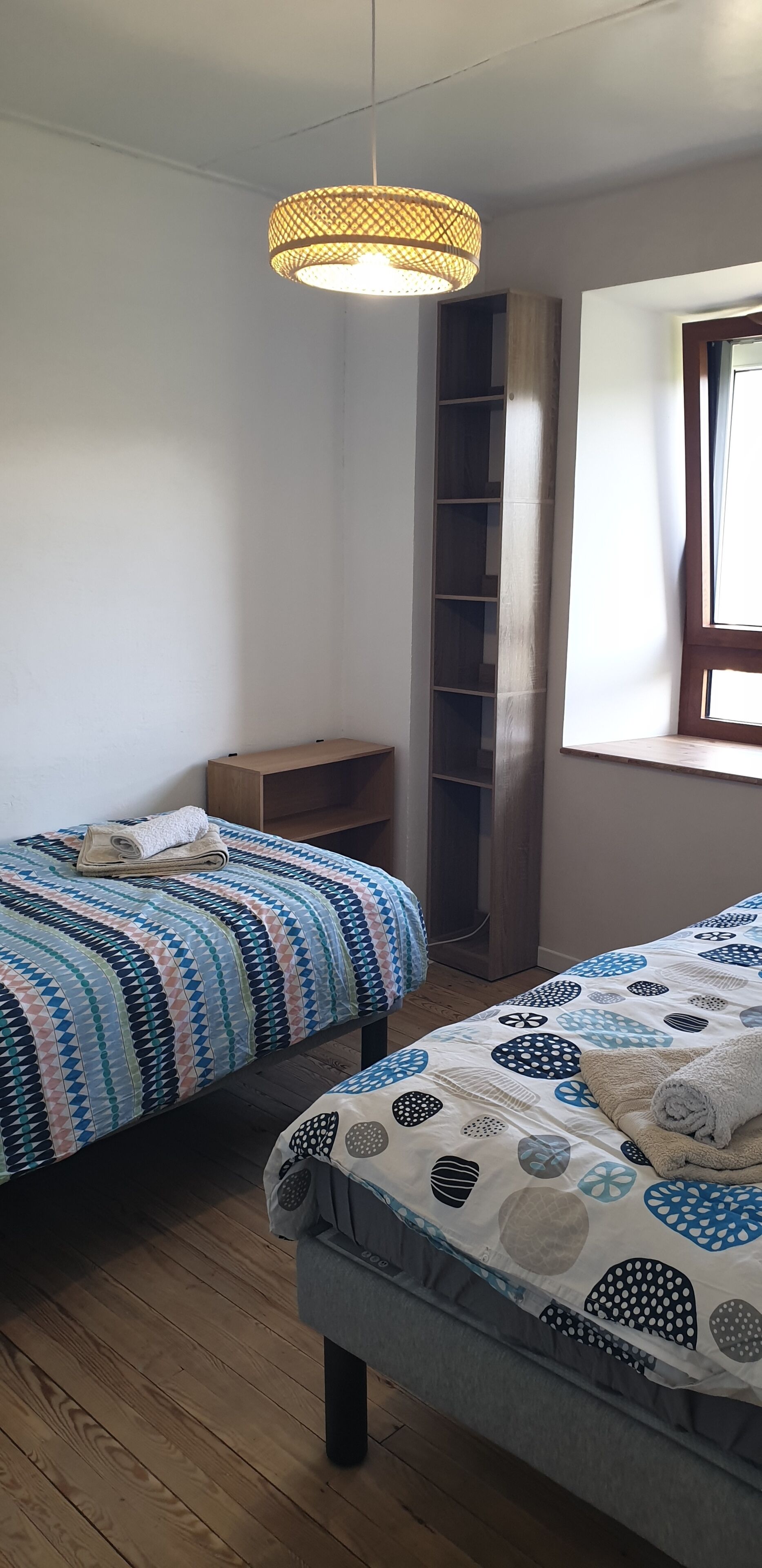 Ein Zimmer mit zwei Betten, einer Hängeleuchte, einem Fenster und einer fest eingebauten Bücherreolen.