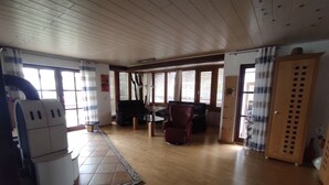 Ein geräumiges Wohnzimmer mit Holzboden, großen Fenstern mit Jalousien, einem Sofa, einem roten Sessel und einem Holzschrank.
