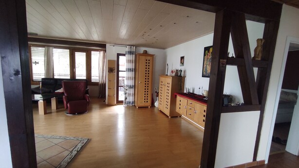 Ein geräumiges Wohnzimmer mit Holzboden, einem roten Sessel, einem Holzschrank und einer Tür zu einem anderen Raum.