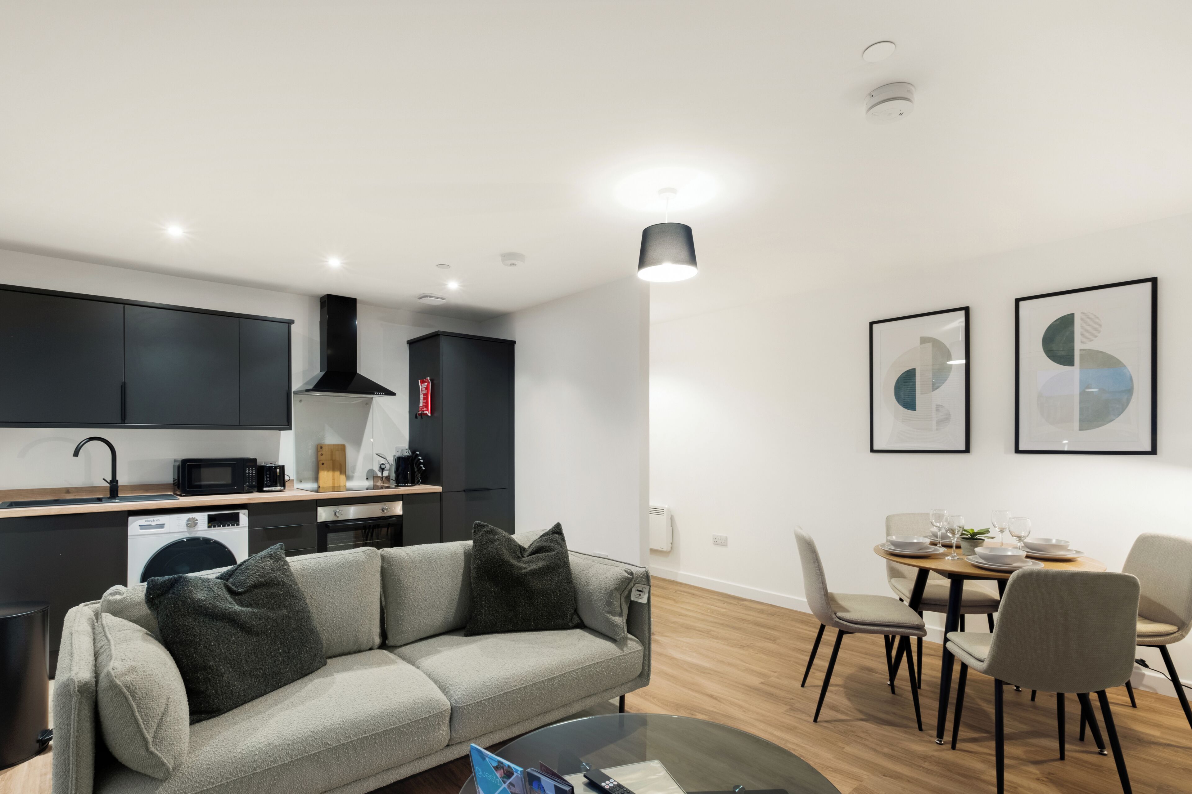 A modern kitchen with dark cabinets, a dining area with a round table and chairs, a gray sofa with cushions, and two framed abstract art pieces on the wall.