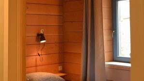 A room with wooden walls, a bed with a pillow, a nightstand, a wall-mounted lamp, a window with curtains, and a door. - Farm Cottage - "Le Perchoir" (Passenans)