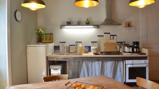 A kitchen with a refrigerator, stove, sink, and dining table with chairs. There are hanging lights, a clock, and various kitchen items on the counter. - Farm Cottage - "Le Perchoir" (Passenans)