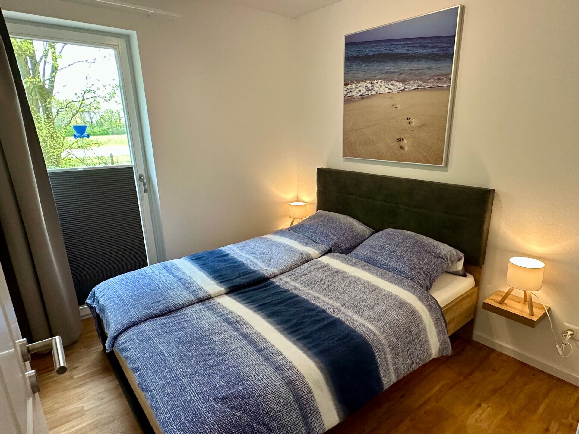 A bedroom with a bed, a nightstand, a wall-mounted lamp, a window, and a beach-themed painting.