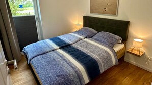 Ett sovrum med en säng, ett nattduksbord, en vägglampa, ett fönster och en tavla med ett strandmotiv.
