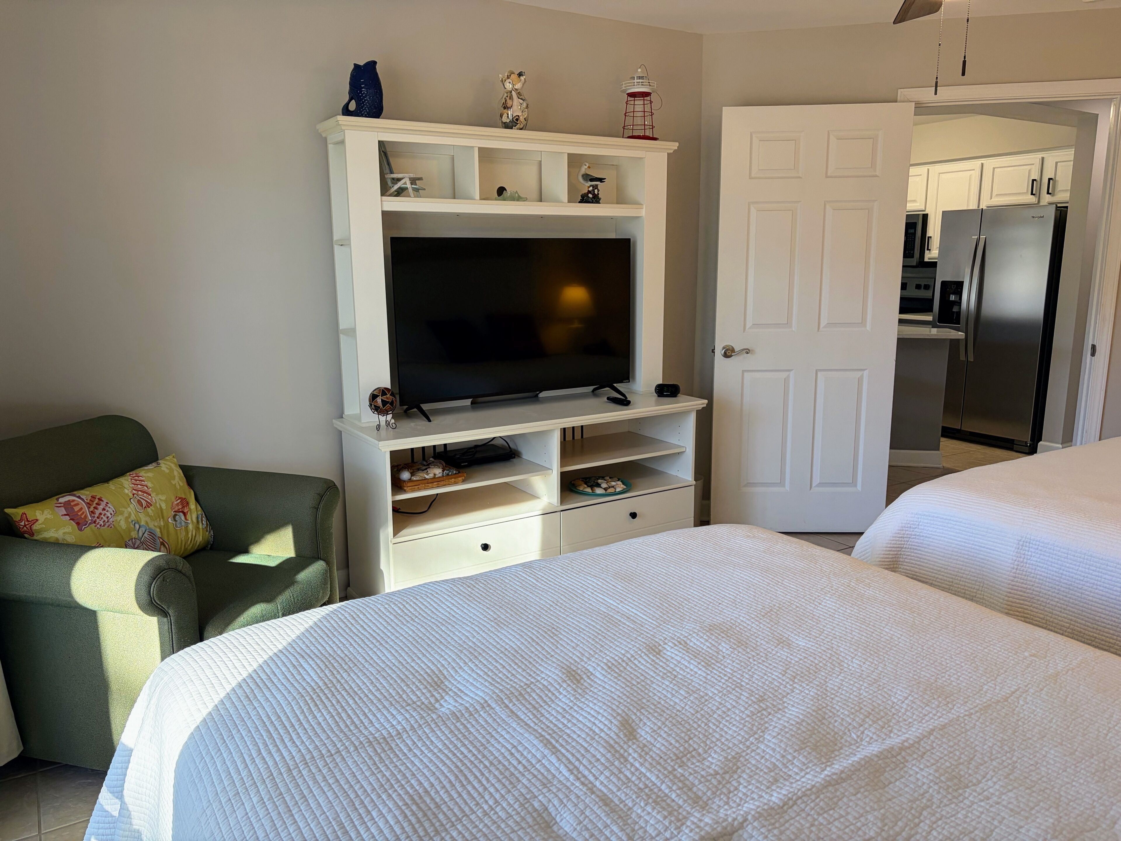 Une pièce avec un téléviseur posé sur un meuble blanc, un fauteuil vert et deux lits avec du linge de lit blanc.