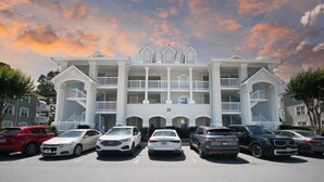 A multi-story building with a central entrance, a parking lot with several cars, and a clear sky with clouds. - Clean and like new condo in Calabash with amazing views of the golf course. (Calabash)