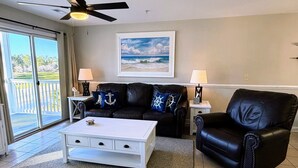 A living room with a white coffee table, two black leather sofas, a ceiling fan, and a painting of a beach scene. - Clean and like new condo in Calabash with amazing views of the golf course. (Calabash)