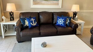 A living room with a brown leather couch, two matching lamps, a framed painting of a beach, and a coffee table. - Clean and like new condo in Calabash with amazing views of the golf course. (Calabash)