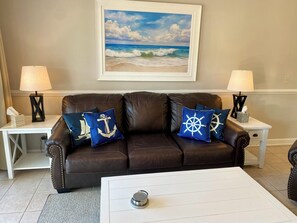 Ein Wohnzimmer mit einer braunen Ledersofa, zwei passenden Lampen, einem gerahmten Gemälde vom Strand und einem Couchtisch.