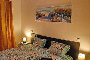 Ein Schlafzimmer mit einem Bett, zwei Kissen, einem Nachttisch mit Lampe, einem Wandbild und einem Vorhang.