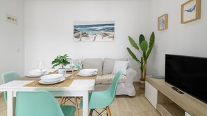 A dining area with a table set for two, a sofa, a TV, and a painting of a beach scene. - Apartment 'Almadraba' with Private Terrace, Wi-Fi and Air Conditioning (Barbate)