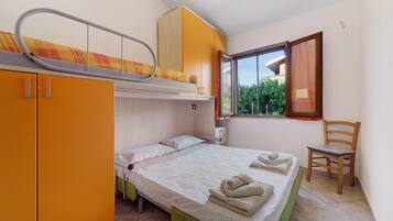A compact room with a bunk bed, a single bed, and a chair. There is a window providing a view of greenery outside.