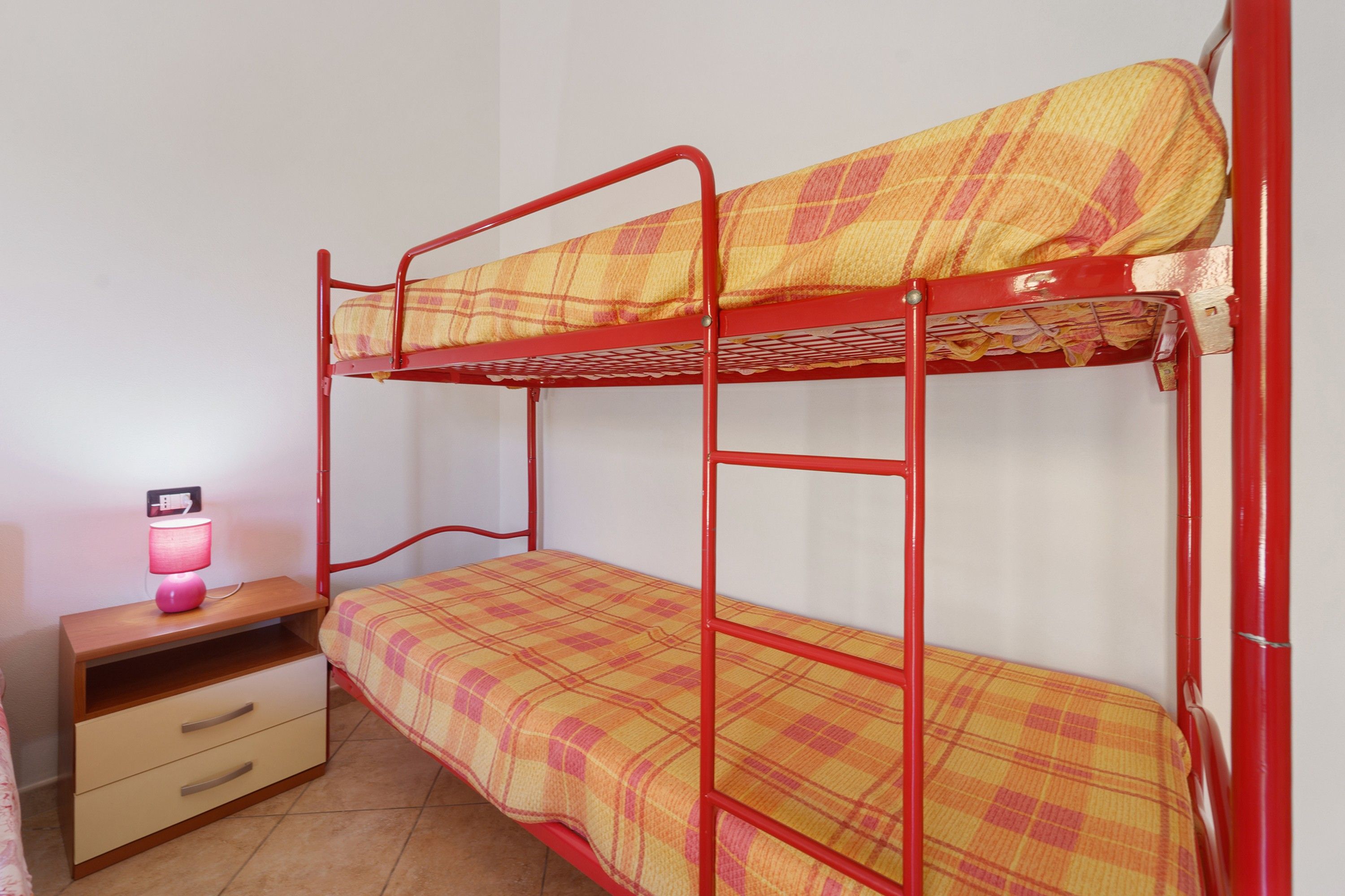 A bunk bed with a plaid bedspread, a bedside table with a lamp, and a drawer unit.