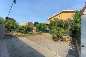 Ein Innenhof mit befestigtem Bereich, ein kleiner Garten mit Sträuchern und Blumen sowie ein Gebäude mit flachem Dach und Satellitenschüssel.