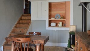 Een trap leidt naar een eetgedeelte met een houten tafel en stoelen, een inbouwkast met keukengerei en een zwarte schoorsteen.