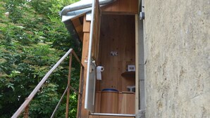 Een houten buitentoilet met een metalen dak, omringd door groen.