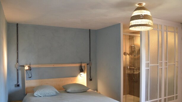 Een slaapkamer met een houten hoofdeinde, twee bedlampen, een bed met kussens en een douchecabine met glazen deur.