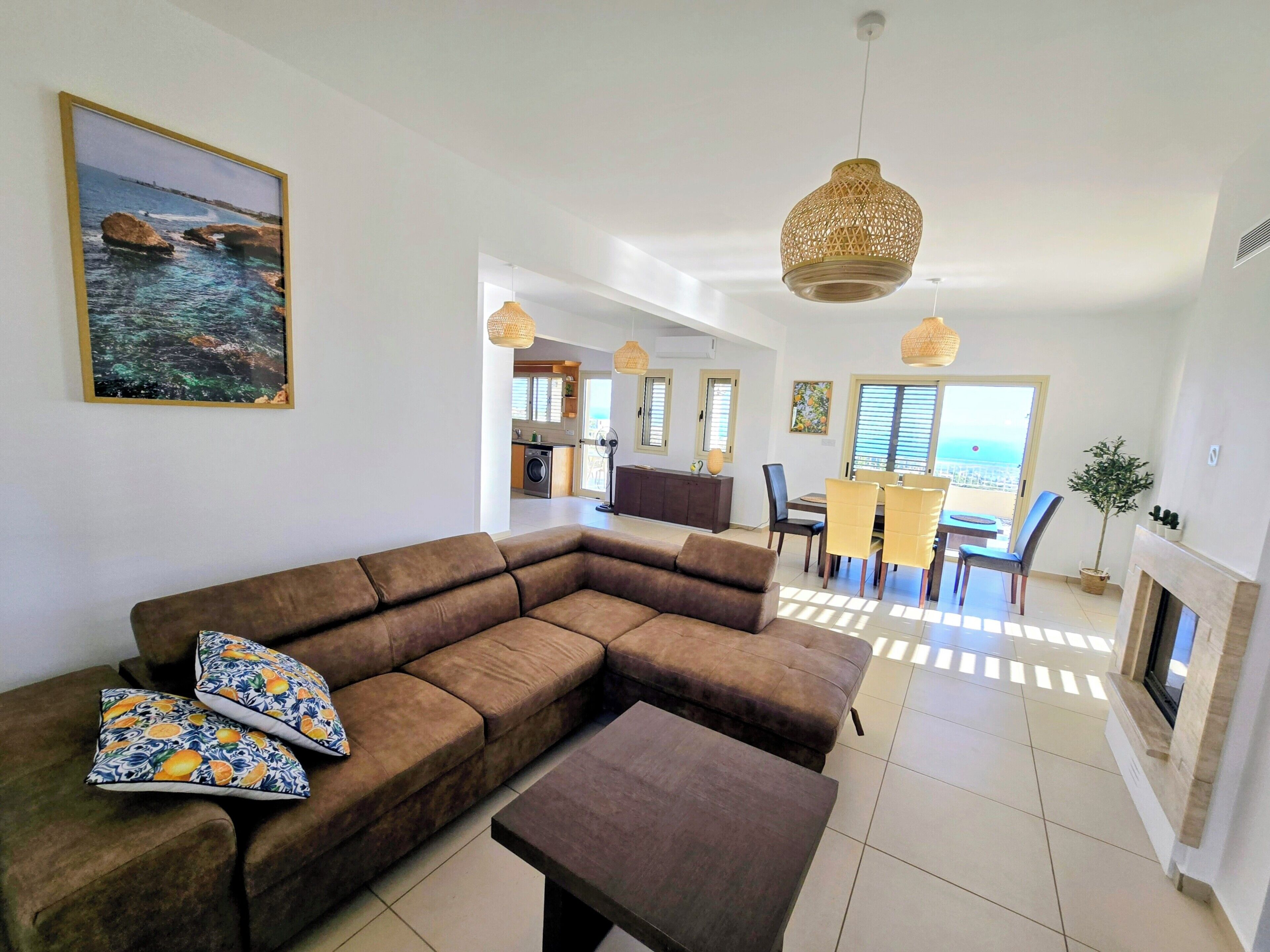 A living room with a sectional sofa, a coffee table, and a dining area with a table and chairs. There is a fireplace and a view of the ocean through the windows.