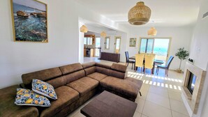 Una sala de estar con un sofá modular, una mesa de centro y un comedor con una mesa y sillas. Hay una chimenea y vistas al océano a través de las ventanas.