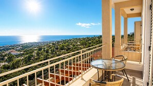 Un balcon doté d’une table et de chaises en verre, offrant une vue sur un paysage côtier avec des maisons et des arbres.