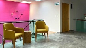 Una zona de recepción con un mostrador de cristal, dos sillas amarillas, una pared rosa y una puerta de madera.
