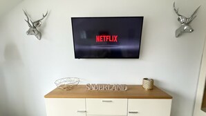 Ett rum med en TV som hänger på väggen och visar "NETFLIX", ett träbord med en dekorativ prydnad märkt "SAUERLAND" och två upphängda hjorthorn på väggen.
