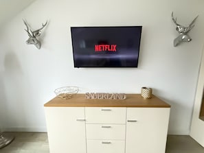 Ein Zimmer mit einem angebrachten Fernseher, der 'NETFLIX' anzeigt, einem Holztisch mit einem dekorativen Gegenstand mit dem Aufdruck 'SAUERLAND' und zwei an der Wand befestigten Hirschgeweihen.