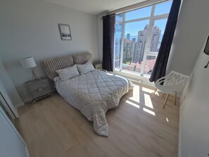 A bedroom with a bed, a chair, a nightstand, a lamp, a window with curtains, and a city view.