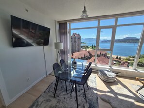 Una camera moderna con un tavolo da pranzo, sedie, una TV montata a parete e un'ampia finestra con vista su un lago e degli edifici.