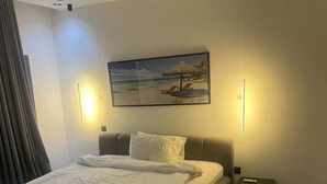 A modern bedroom with a large bed, a nightstand, a painting of a beach scene, and a ceiling light. - JOYZY HAVEN LUXURY SERVICE APARTMENT  (Ibadan)