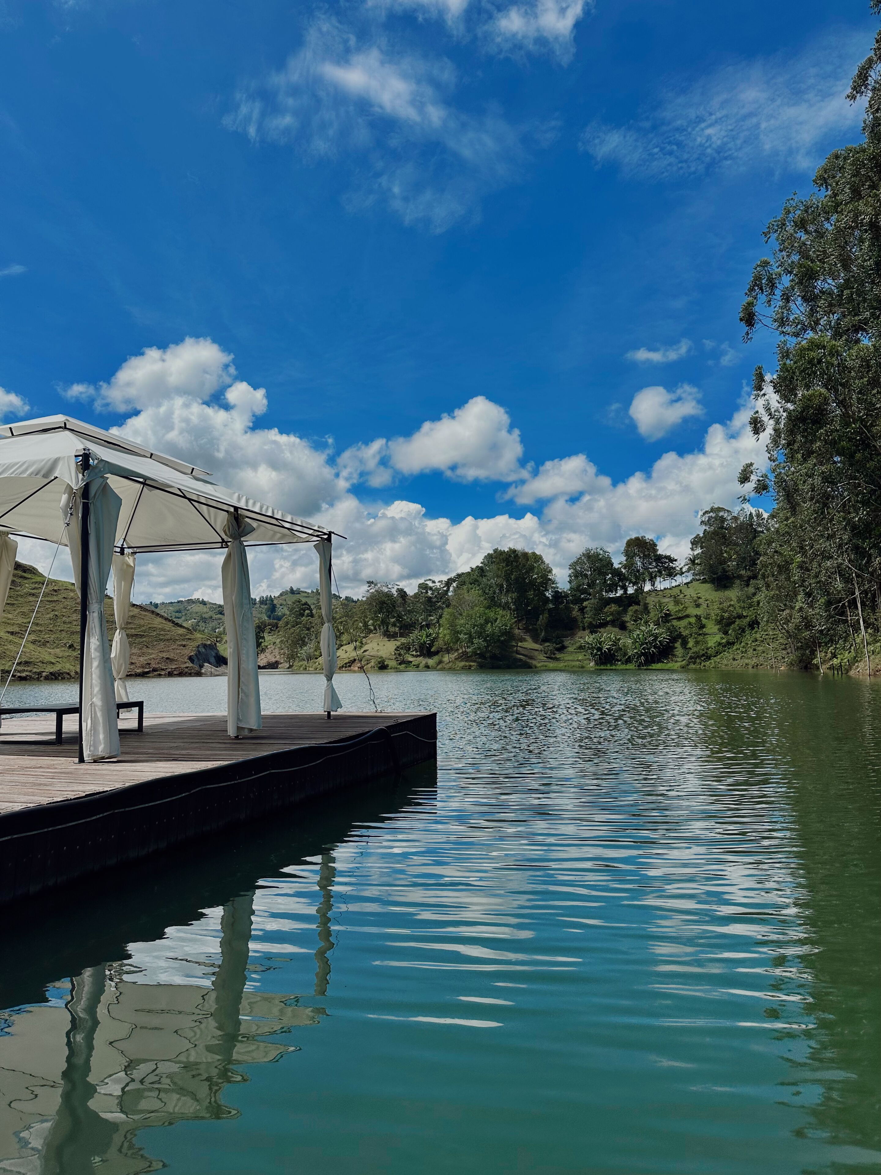 Cenário tranquilo à beira do lago, com um píer de madeira e um pavilhão coberto.