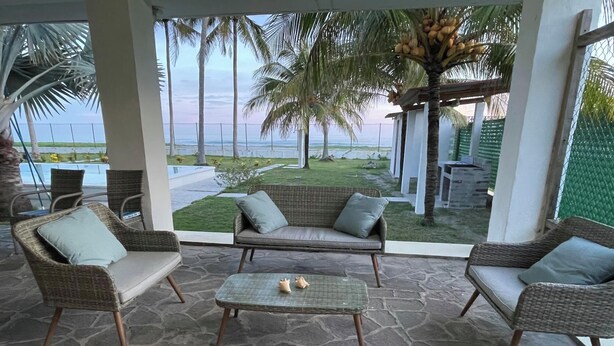 A covered outdoor seating area with wicker furniture and a stone floor, overlooking a grassy lawn and palm trees. - Enjoyable 4-bedroom country house with AC in Playa El Amatal (playa el amatal)