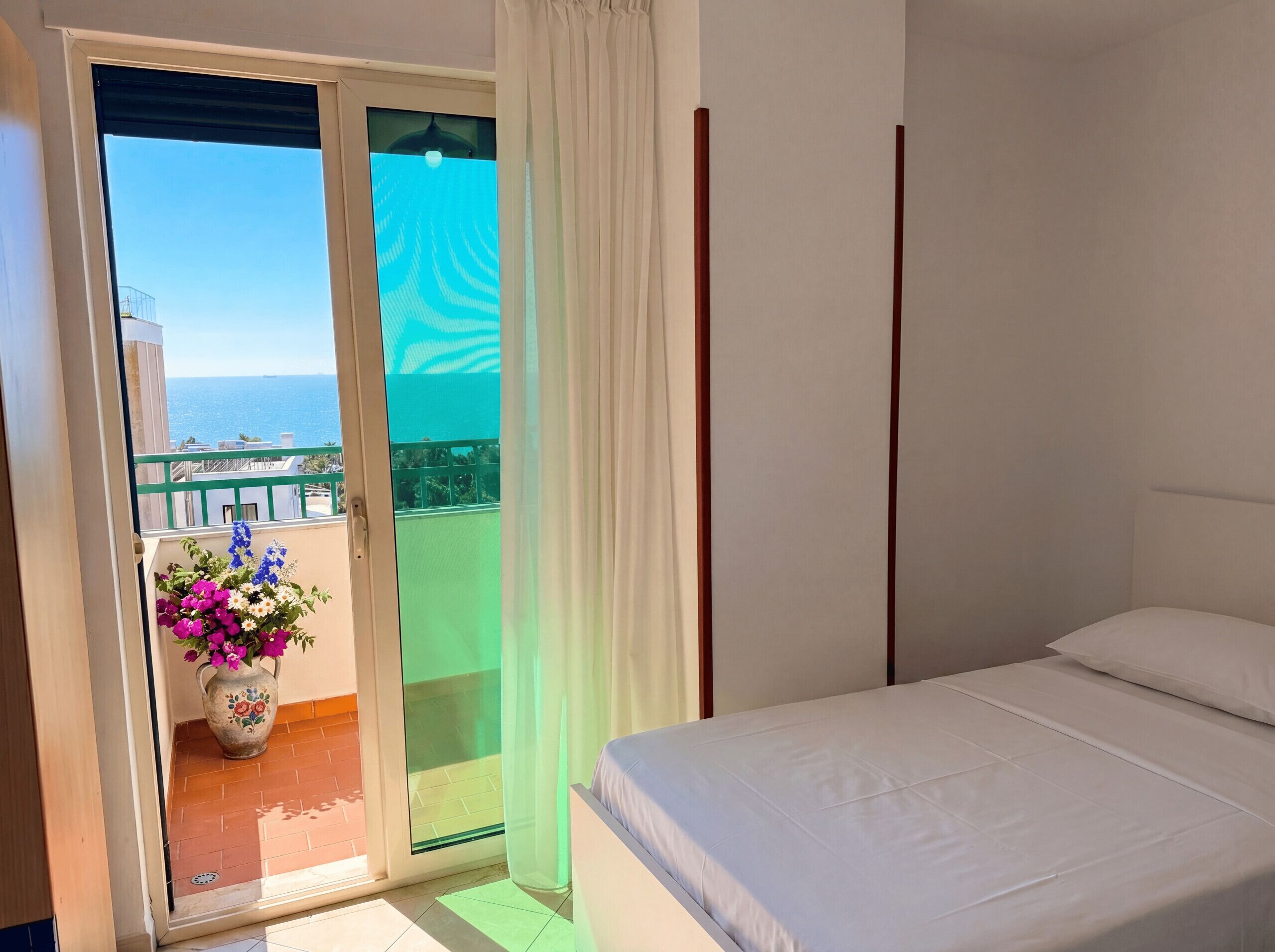 Quarto com cama, vista para o mar e varanda com um vaso de flores.