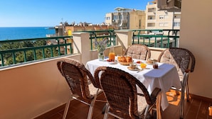 A balcony with wicker chairs, a table set for breakfast, and a view of the sea and buildings. - Holiday Home by the Beach Durres 117 (Durres)
