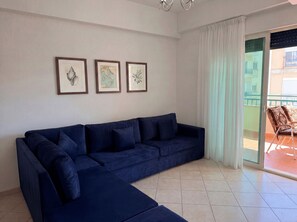 A living room with a dark blue sofa, three framed pictures on the wall, and a sliding glass door leading to an outdoor balcony. - Holiday Home by the Beach Durres 117 (Durres)
