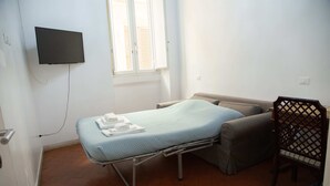 Une chambre avec un lit simple, un téléviseur fixé au mur, une fenêtre avec des stores, une table de chevet et une chaise.