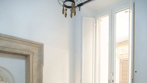 Une chambre à coucher avec un lit, une fenêtre avec des volets, une lampe fixée au mur et un lustre.