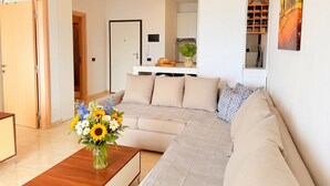 A modern living room with a sofa, a wooden coffee table with a vase of flowers, a kitchen area in the background, and a painting on the wall. - Beachfront- Sea View Holiday Home 105 (Durres)