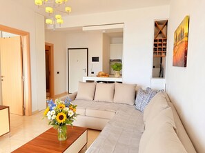 Un salon moderne avec un canapé, une table basse en bois ornée d’un vase de fleurs, un coin cuisine à l’arrière-plan et un tableau accroché au mur.
