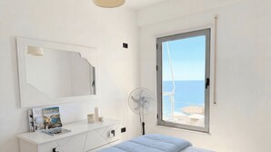 A bedroom with a bed, a bedside table with a lamp, a fan, and a window with a view of the sea and wind turbines. - Beachfront- Sea View Holiday Home 105 (Durres)