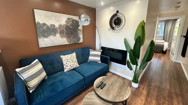 Una sala de estar con un sofá azul, una mesa de centro redonda de madera, un televisor montado y una planta verde grande.
